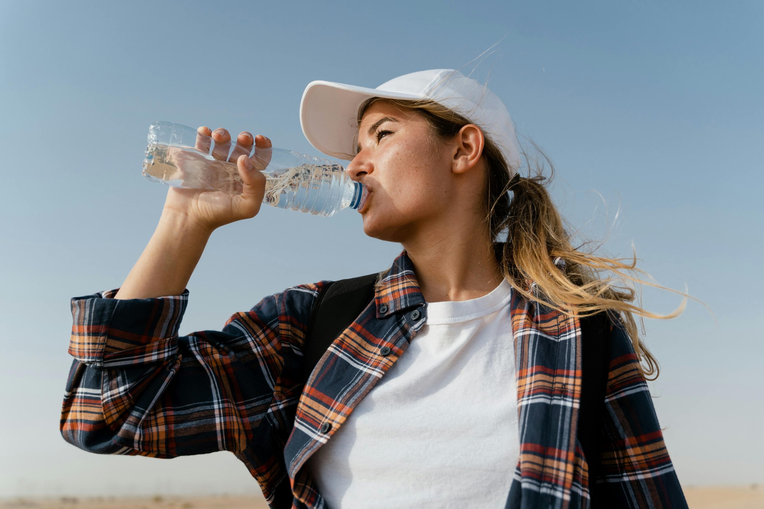 mujer de pelo rubio bebiendo agua de una botella con remera blanca, arriba una camisa abierta a cuadros y un gorro blanco.