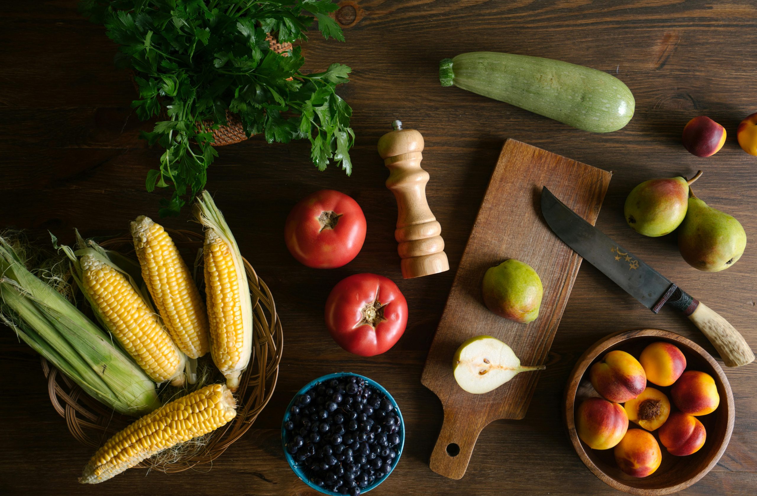 se observa una tabla con frutas y verduras saludables con una cuchilla prontas para ser cortadas. También se ve un molinillo de pimienta