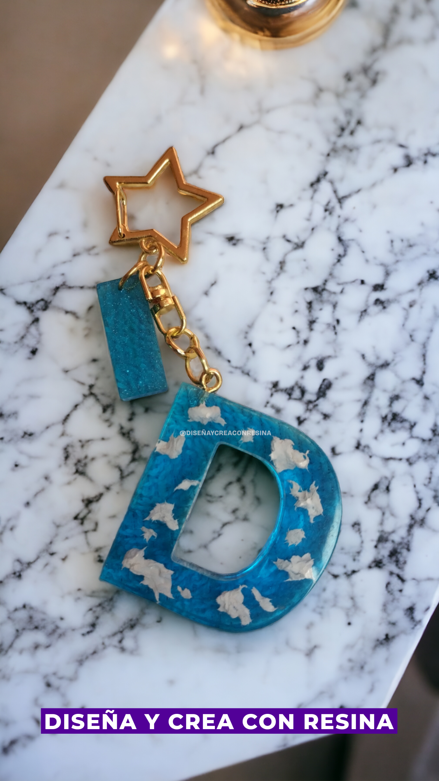 llavero dorado con una estrella chica del cual cuelga una letra D mayúscula de color azul con manchas blancas y una barra pequeña también del mismo color azul que la letra D pero sin las manchas