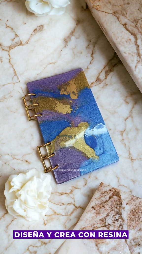 En la imagen se observa lo que sería una tapa de agenda hecha con resina epóxica, con dos espirales de color dorado que sujetan la tapa, y esta tapa es una mezcla de colores: violeta, azul dorado y celeste.