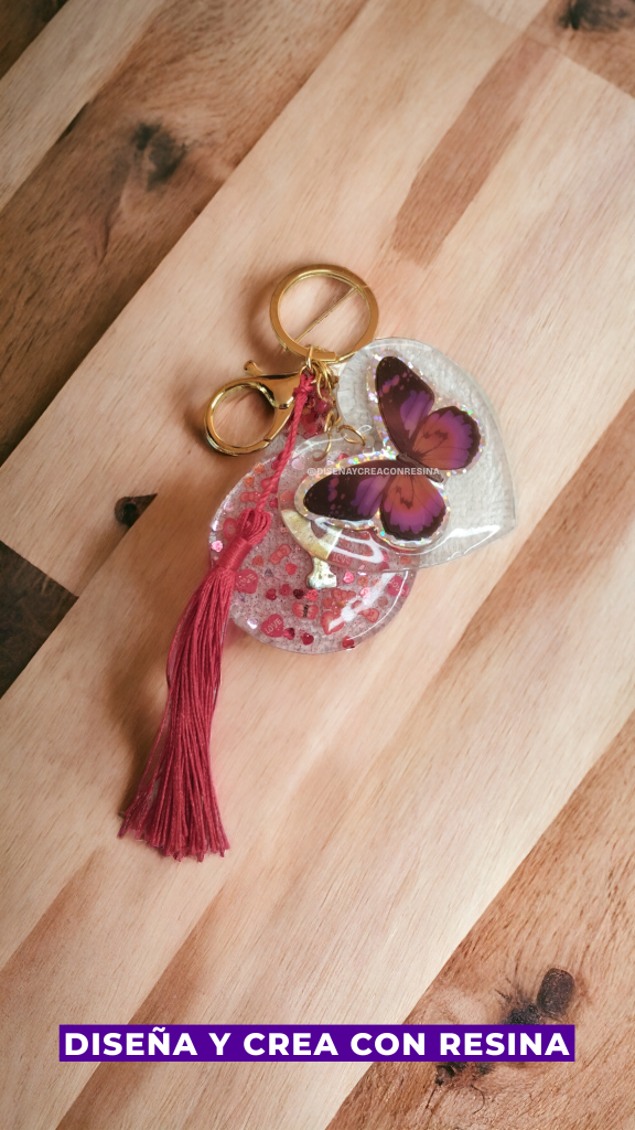En esta imagen se muestra un llavero doble realizado con resina epóxica, sobre una mesa de madera clara uno que es un corazón transparente con una mariposa en el centro con clores dentro de la tonalidad del morado, el otro es un círculo transparente con puntitios rosados y haciendo juego un hilo del cual cuelga como una cola de caballo de hilos del mismo color.