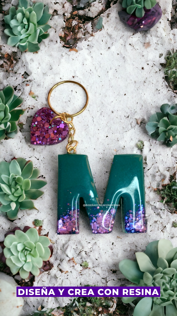 En esta imagen se muestra un llavero doble realizado con resina epóxica, uno que es un corazón brillante de color violeta y el otro es una letra M mayúscula con color verde oscuro arriba y abajo una tonalidad de violeta claro. También en la imagen se observa que el llavero está apoyado sobre un suelo de arena con tunas alrededor