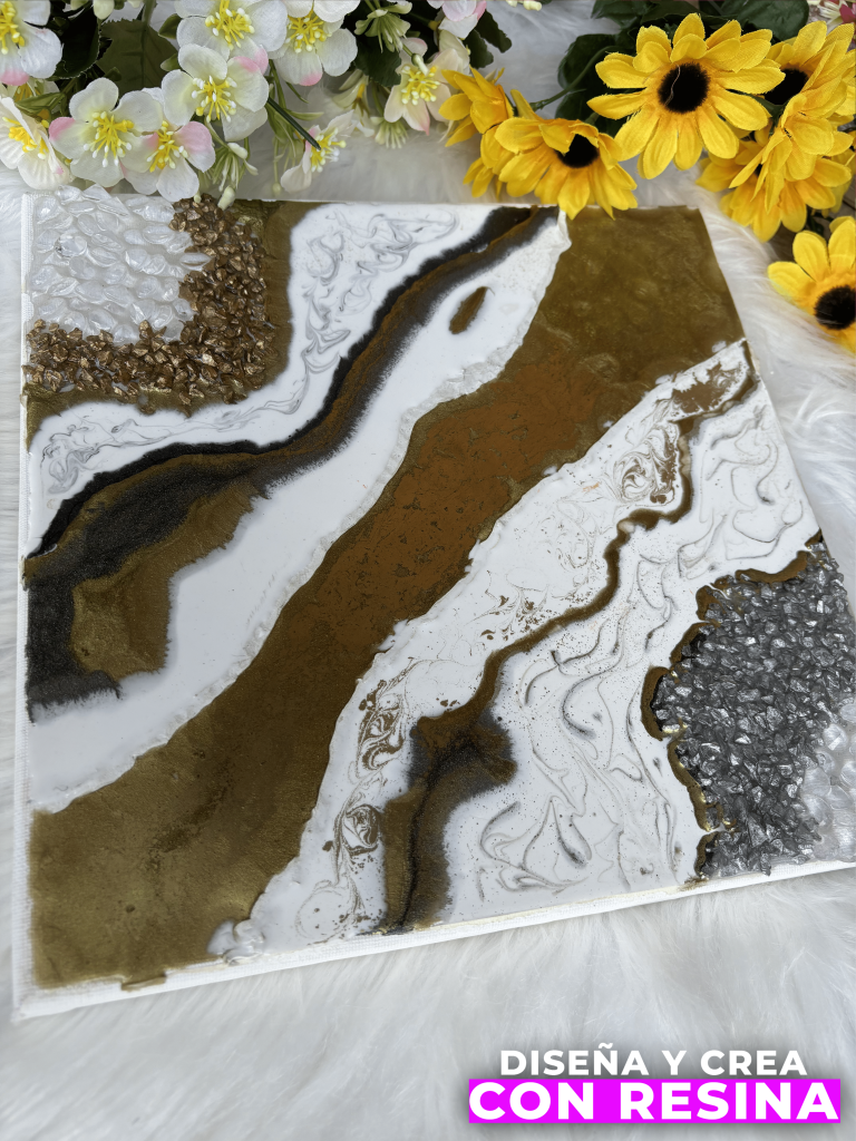 En la imagen vemos un cuadro realizado con resina epóxica con colores que van de forma diagonal de un margen al otro del cuadro, intercalando colores blanco, blanco con líneas negras, marrón en diferentes tonos y gris brillante. En la parte superior se observan flores que están por fuera del cuadro, amarillas y otras blancas. 