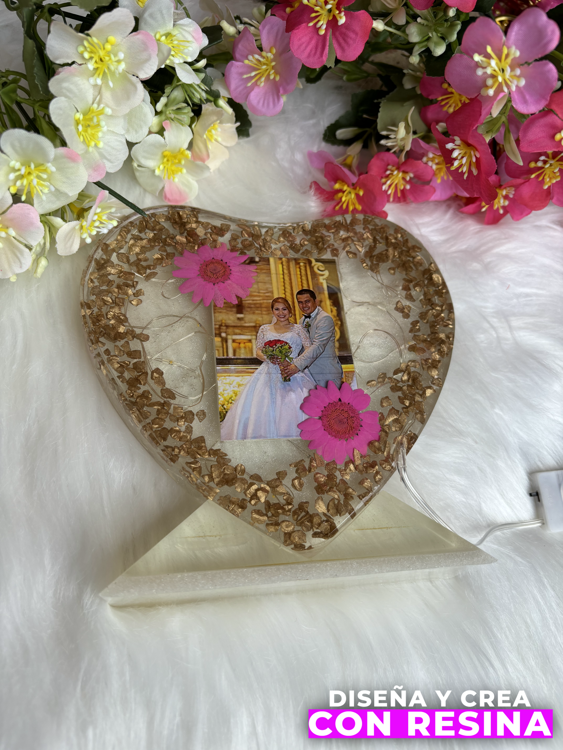 lampara con forma de corazón transparente, en el medio una imágen de bodas, con dos flores rosadas (una en la esquina superior izquierda, la otra en la esquina inferior derecha). sobre una superficie blanca