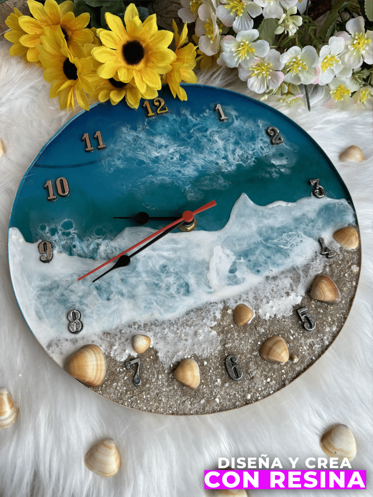 En la imagen se muestra un reloj realizado con resina epóxica con la temática de una playa donde se ve la arena, cucharitas de playa y el océano con espuma de mar. En la parte superior se observan dos tipos de flores, unas de color amarillo y otras de color blanco