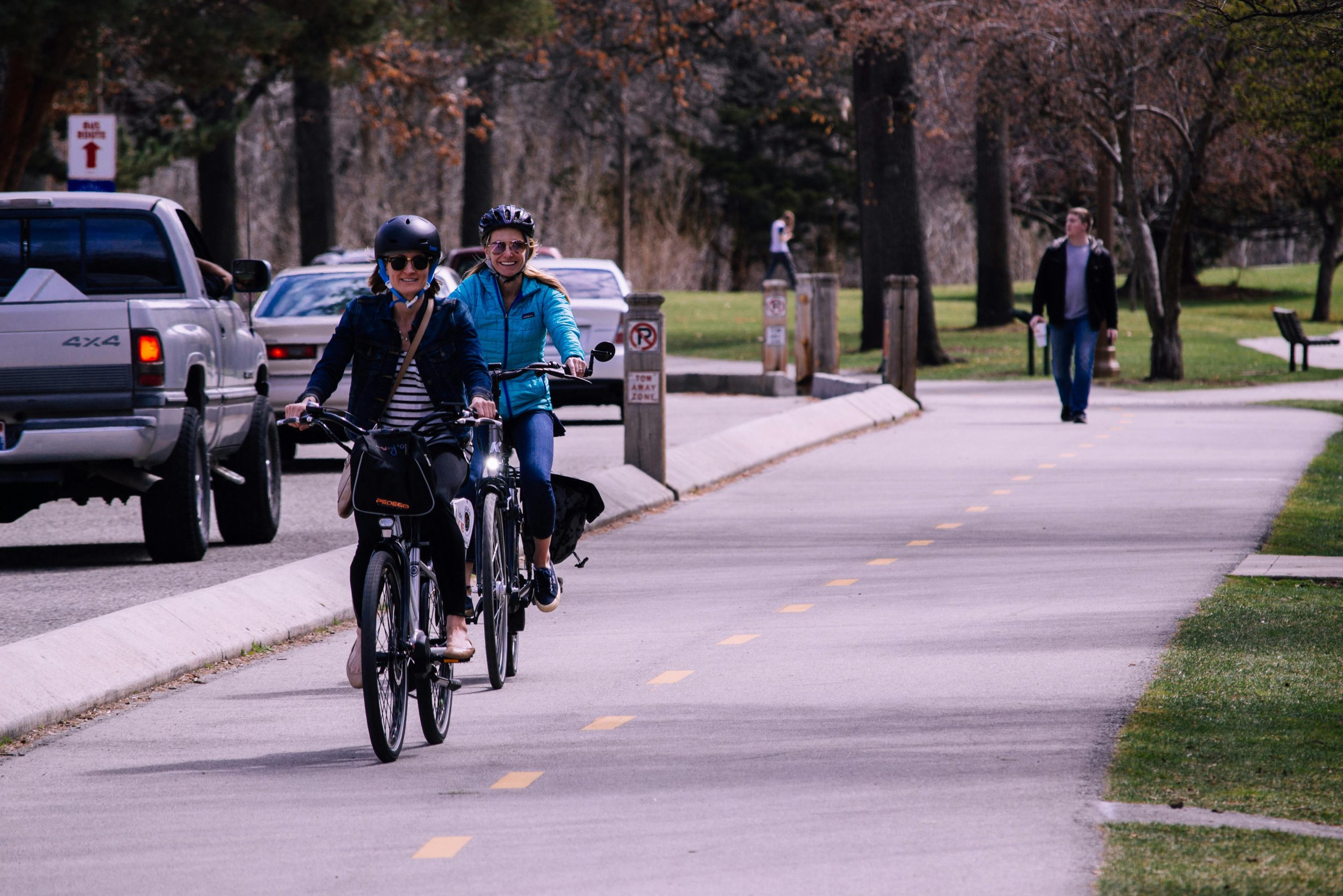 2 personas en un día normal trasladándose en bicicleta por un parque y a lo lejos se ve una persona que viene caminando, al costado se ve la calle y vehiculos