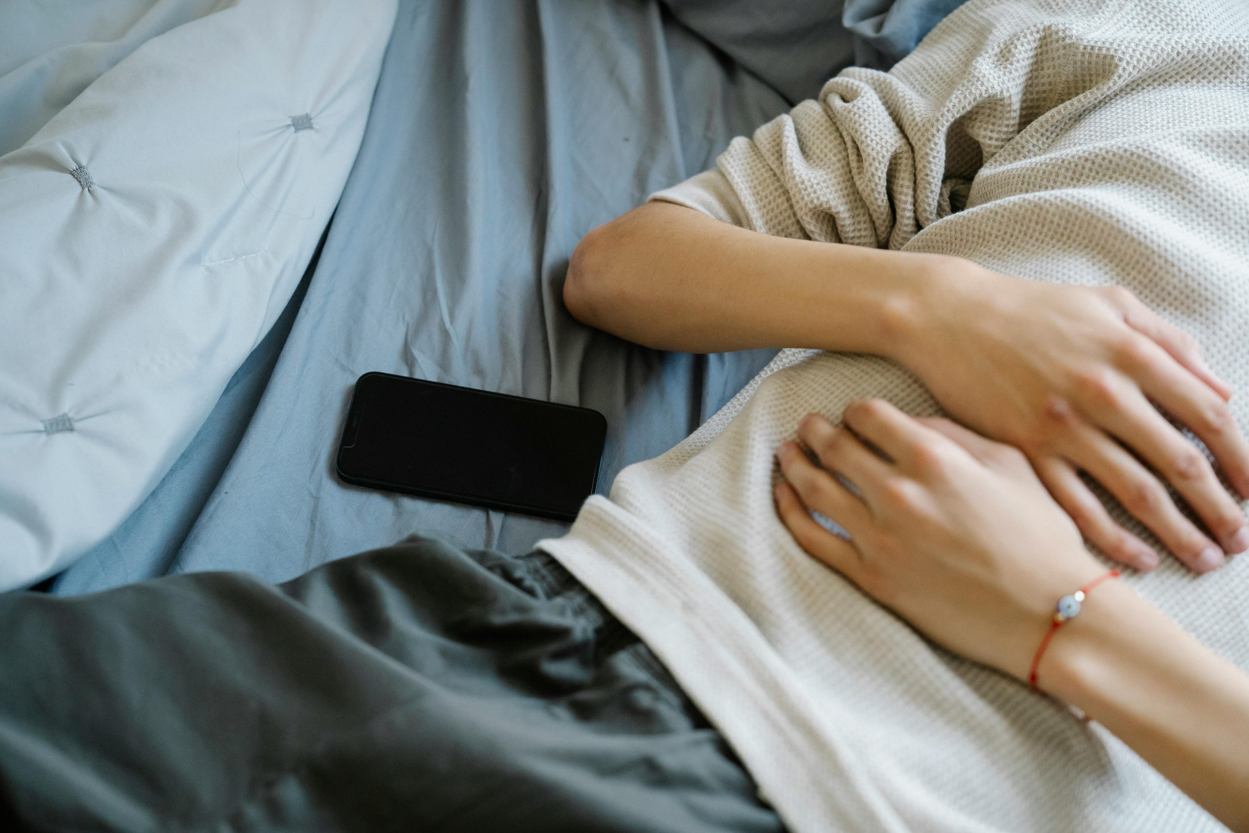 se observa persona durmiendo sobre una cama donde sólo pueden verse sus brazos sobre su abdomen y apenas una parte del pantalon, con el celular apagado a su lado