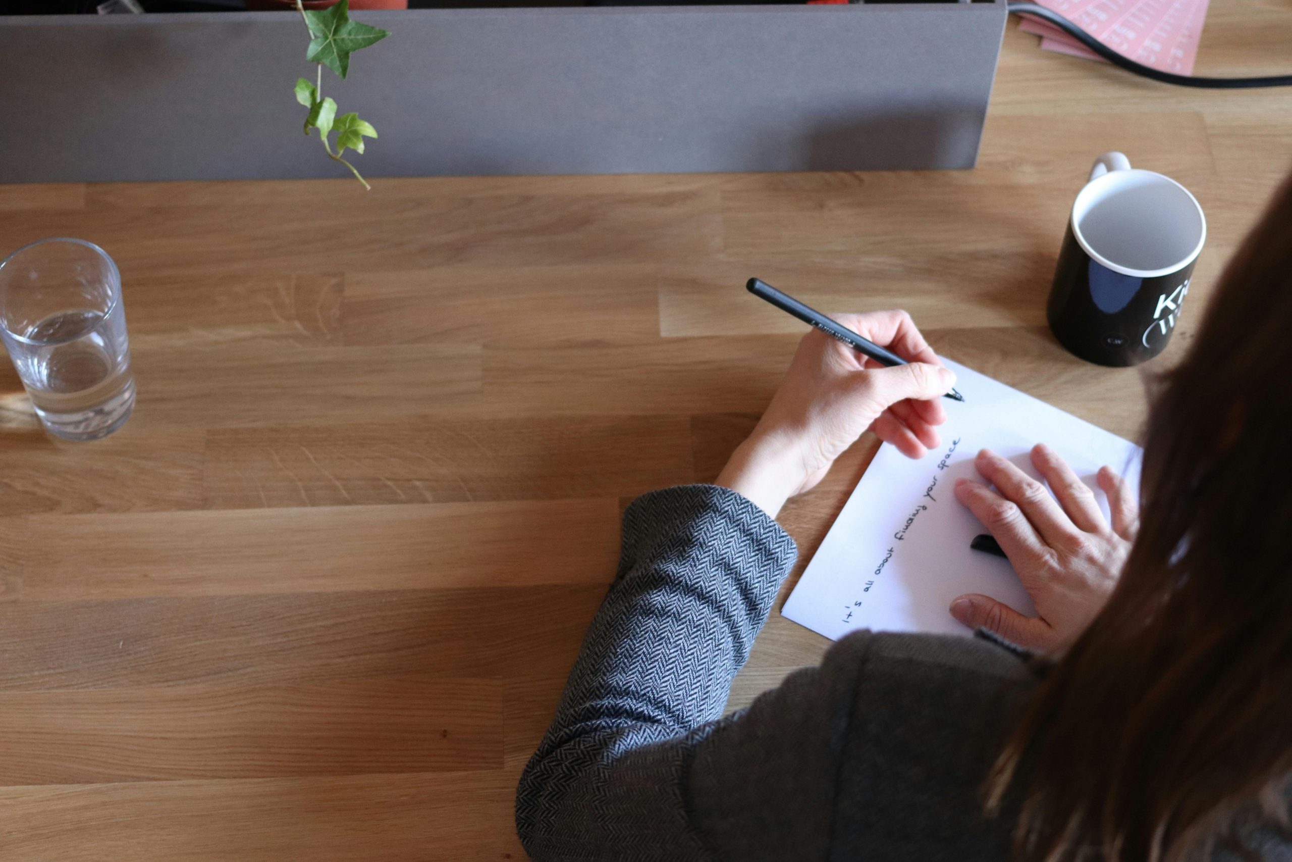 Una persona sentada en un escritorio minimalista, con luz natural entrando por una ventana, escribiendo en un cuaderno. Al lado: un vaso de agua y una taza de café (sin protagonismo). Fondo limpio, tonos cálidos, sensación de mañana tranquila.