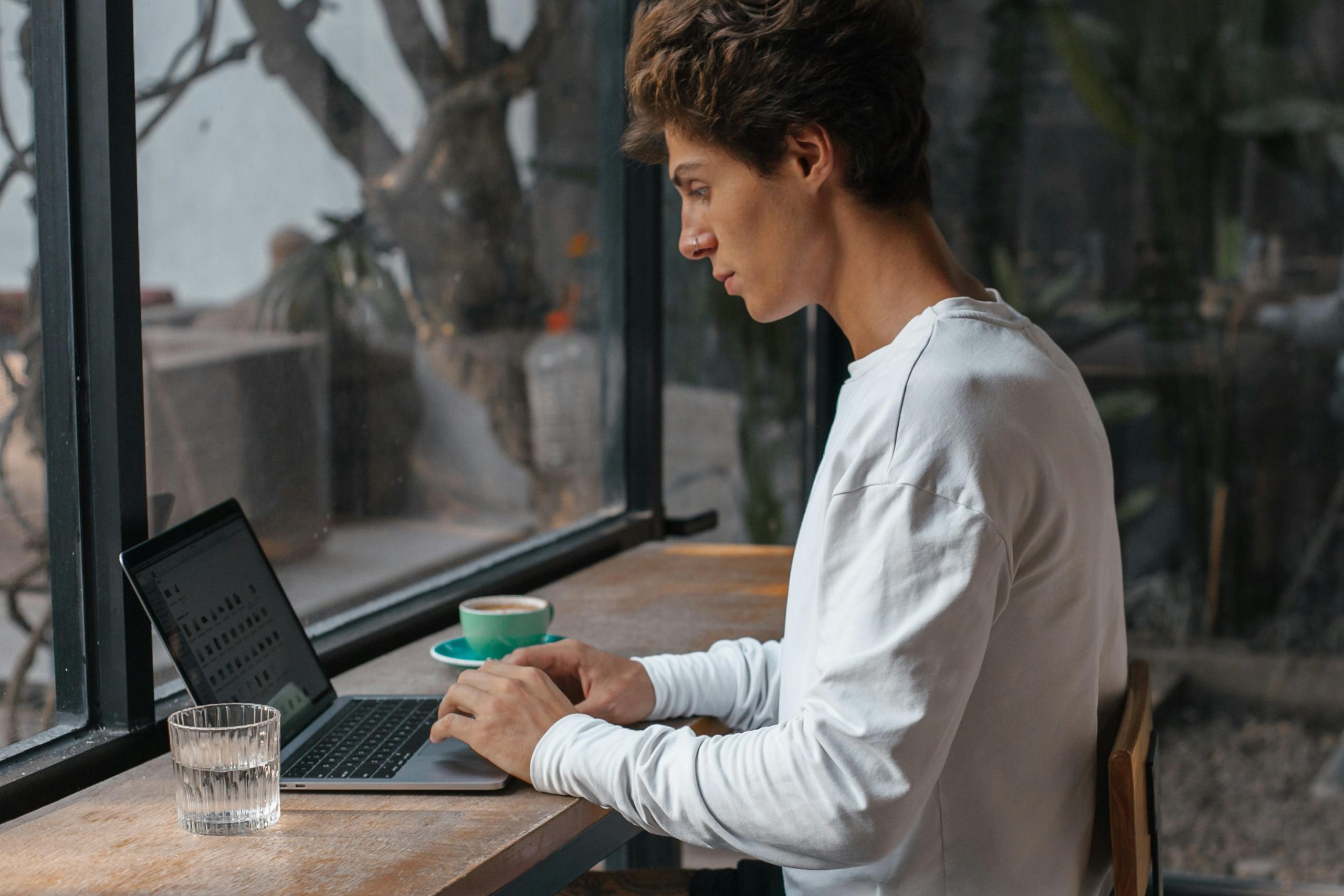 Una persona trabajando concentrada frente a una laptop en un espacio minimalista, con luz natural entrando por una ventana. un vaso de agua y una taza de café